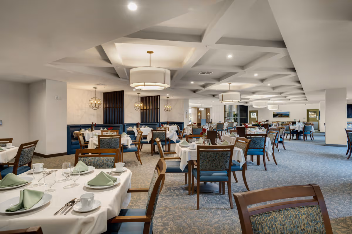 Large, elegant dining room with neatly set tables, upholstered wooden chairs, and pendant lights under a coffered ceiling.