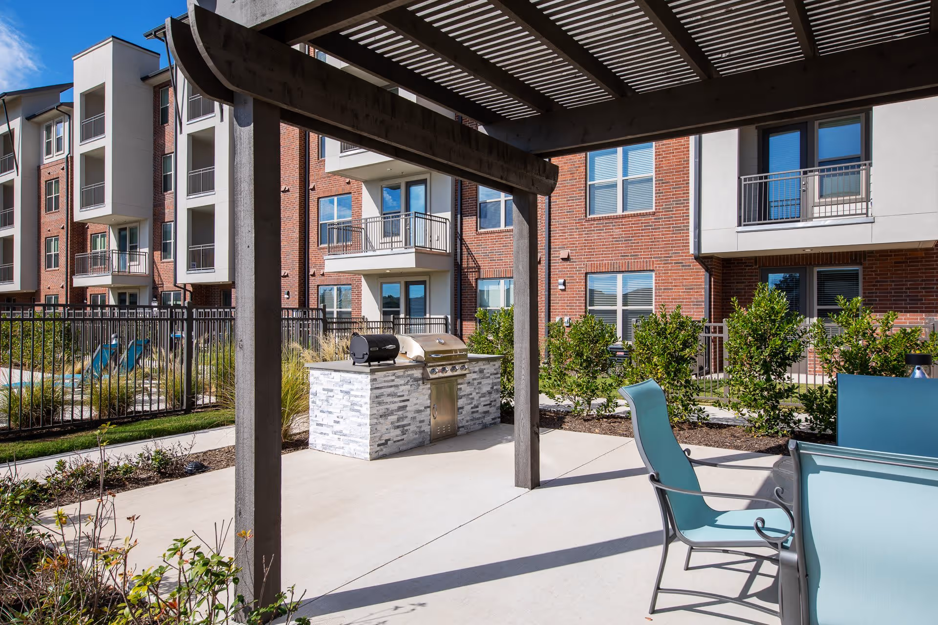 Outdoor patio area with a pergola providing shade over a seating area with blue chairs and a table. In the background, there is a built-in grill station with a stainless steel grill and a smoker. Surrounding the patio are shrubs and a multi-story brick apartment building with balconies and windows.