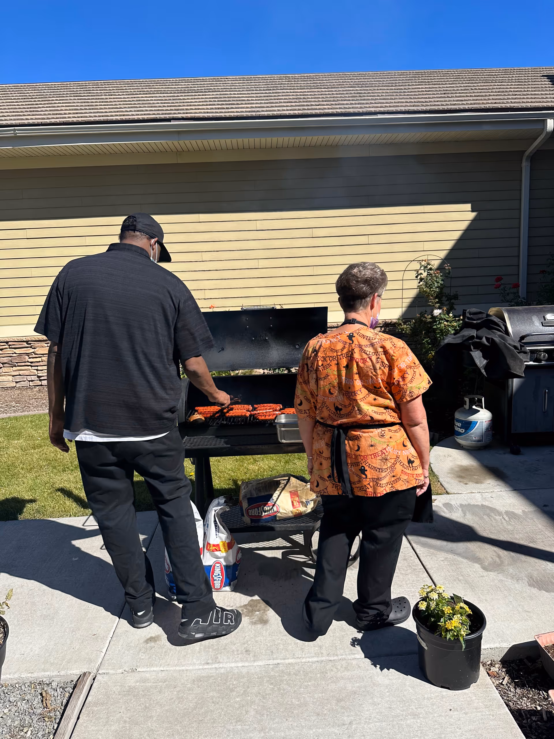 Two people standing outdoors next to a barbecue grill with sausages cooking on it. One person is wearing a black shirt, black pants, and a black cap, while the other person is wearing an orange patterned shirt and black pants. There are bags of charcoal and a propane tank nearby, with a building wall and some plants in the background under a clear blue sky.