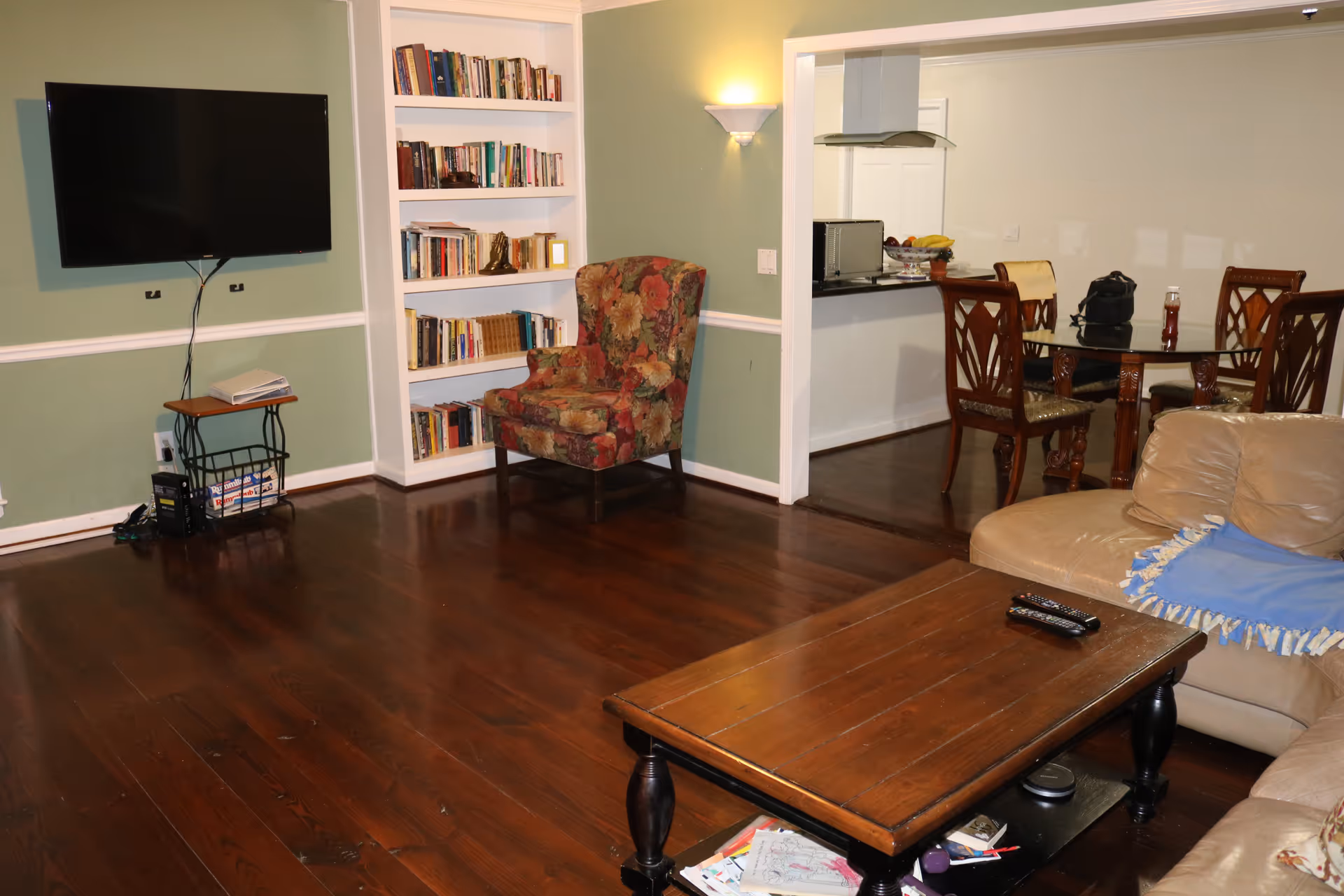 A cozy living room with a wall-mounted TV, built-in bookshelf, floral armchair, coffee table and a view into a dining area.