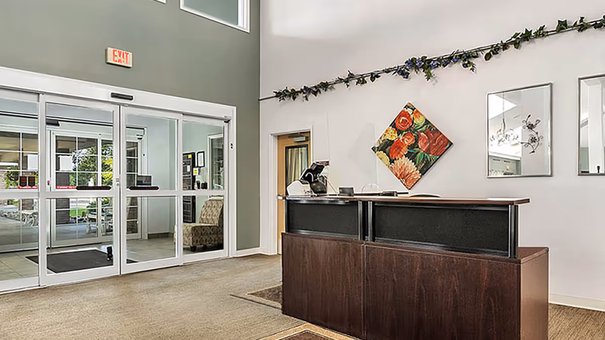 Reception area of Holiday Genesee Gardens facility with a dark wood front desk, floral artwork on the wall, mirrors, and a garland decoration above. Glass automatic sliding doors lead to an outside area with seating visible through the glass.