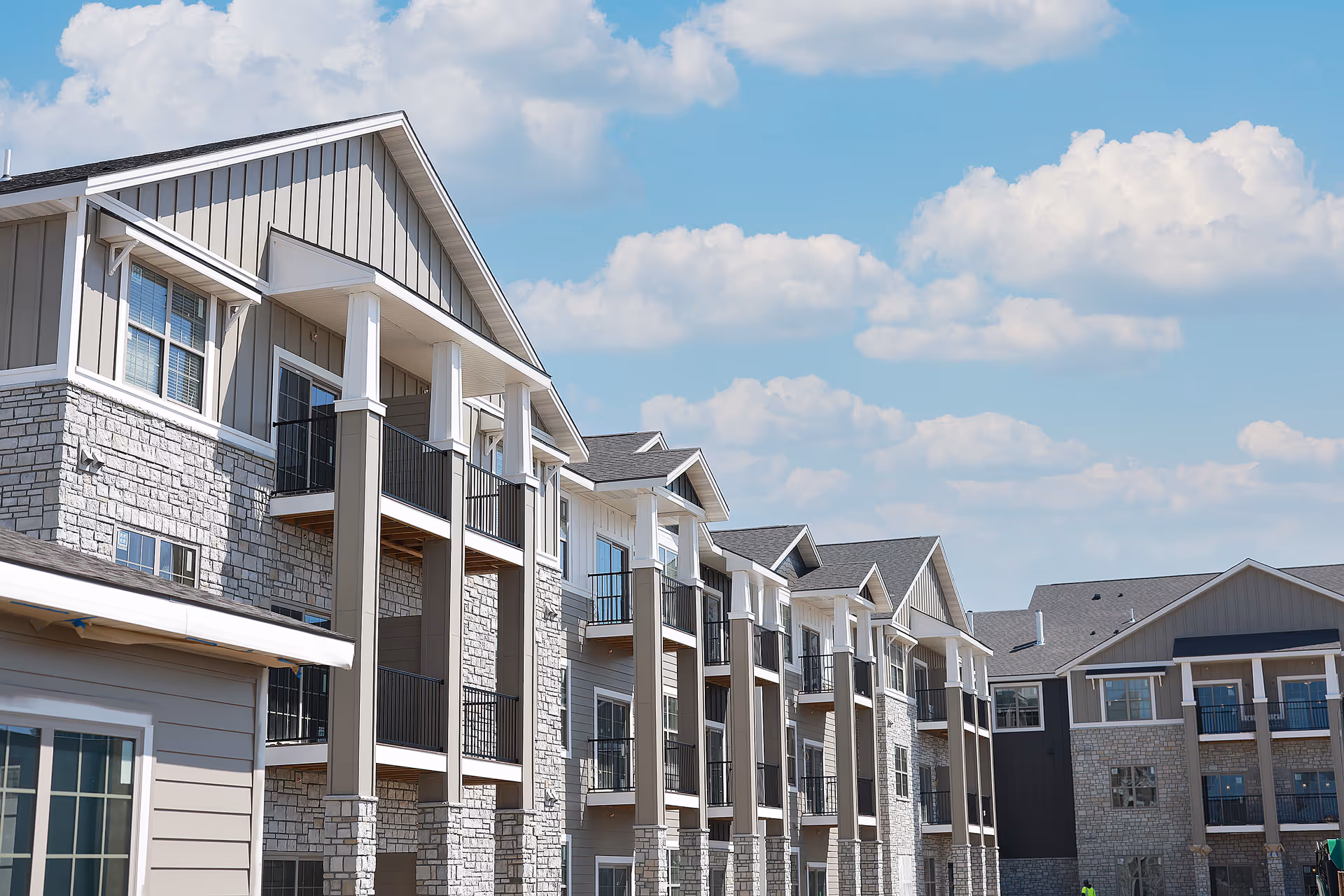 Exterior view of a multi-story senior living facility building with balconies, stone and siding facade, under a partly cloudy blue sky.