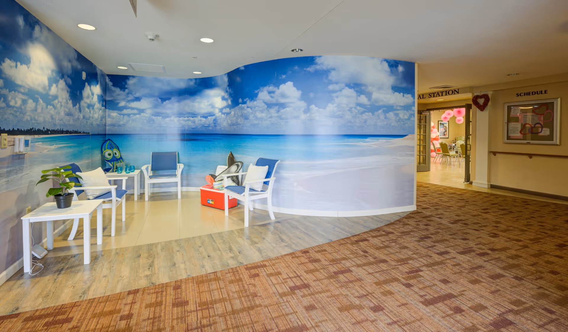 A seating area with white chairs and small tables is set against a wall mural depicting a bright beach scene with blue sky, clouds, and ocean. The floor transitions from wood to carpet, and a hallway with a bulletin board and schedule is visible in the background.