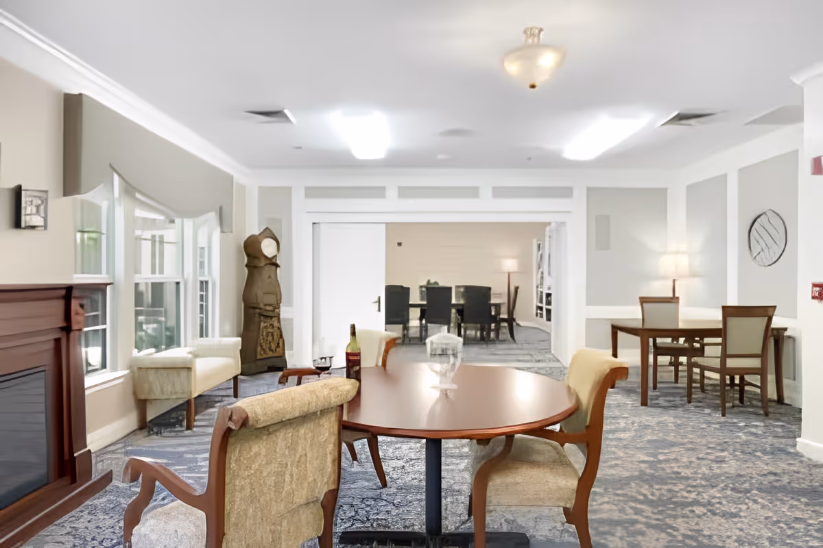 Well-lit communal dining and lounge area with round tables, upholstered chairs, a fireplace and bay windows.