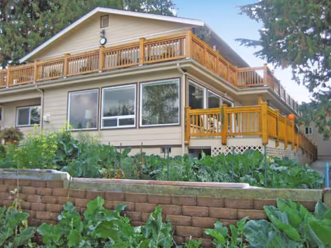 Two-story beige house with wooden balconies and a terraced garden in the foreground.