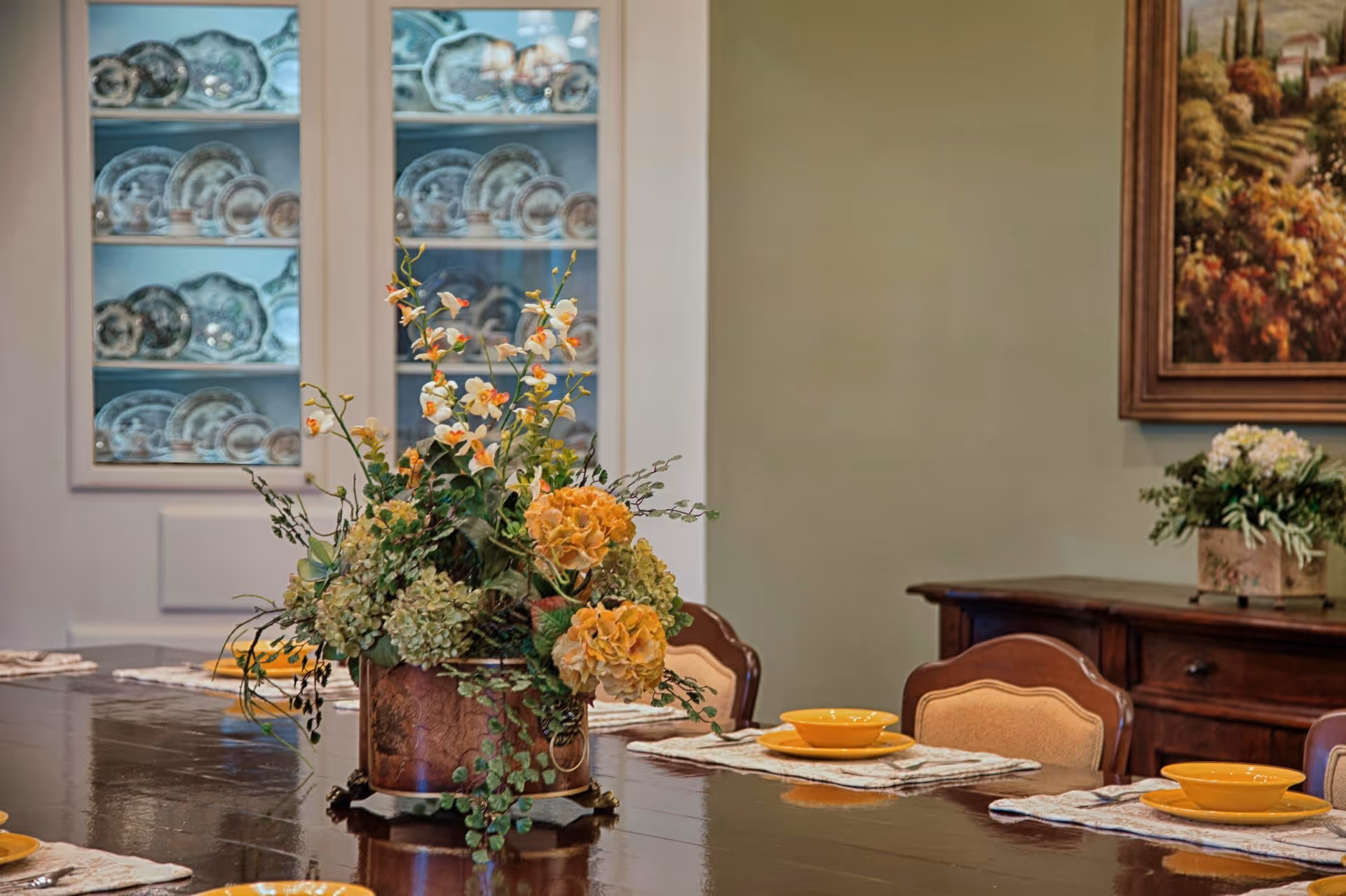 A dining room with a polished wooden table set with yellow bowls and placemats. A large floral arrangement in a decorative vase is centered on the table. In the background, there is a cabinet with glass doors displaying decorative plates and a sideboard with a smaller floral arrangement and a framed painting on the wall.