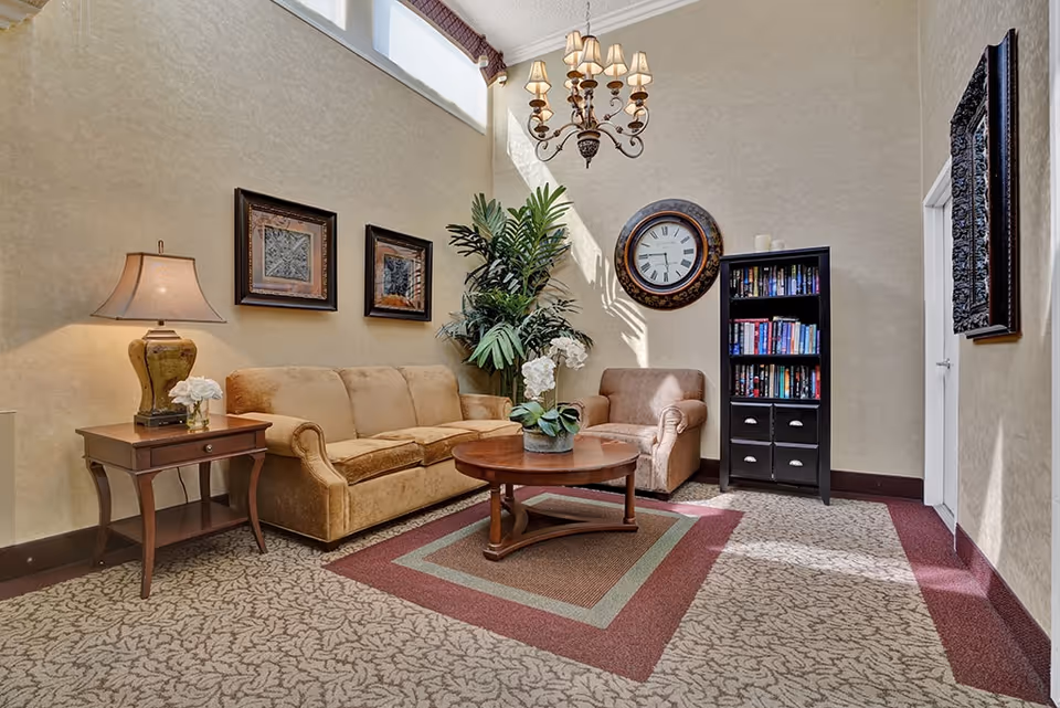 A cozy living room area with a beige sofa, a matching armchair, a wooden coffee table with a flower arrangement, a side table with a lamp and flowers, a tall black bookshelf filled with books, a large decorative wall clock, two framed artworks on the wall, and a large plant. The room has high ceilings with windows near the top letting in natural light.