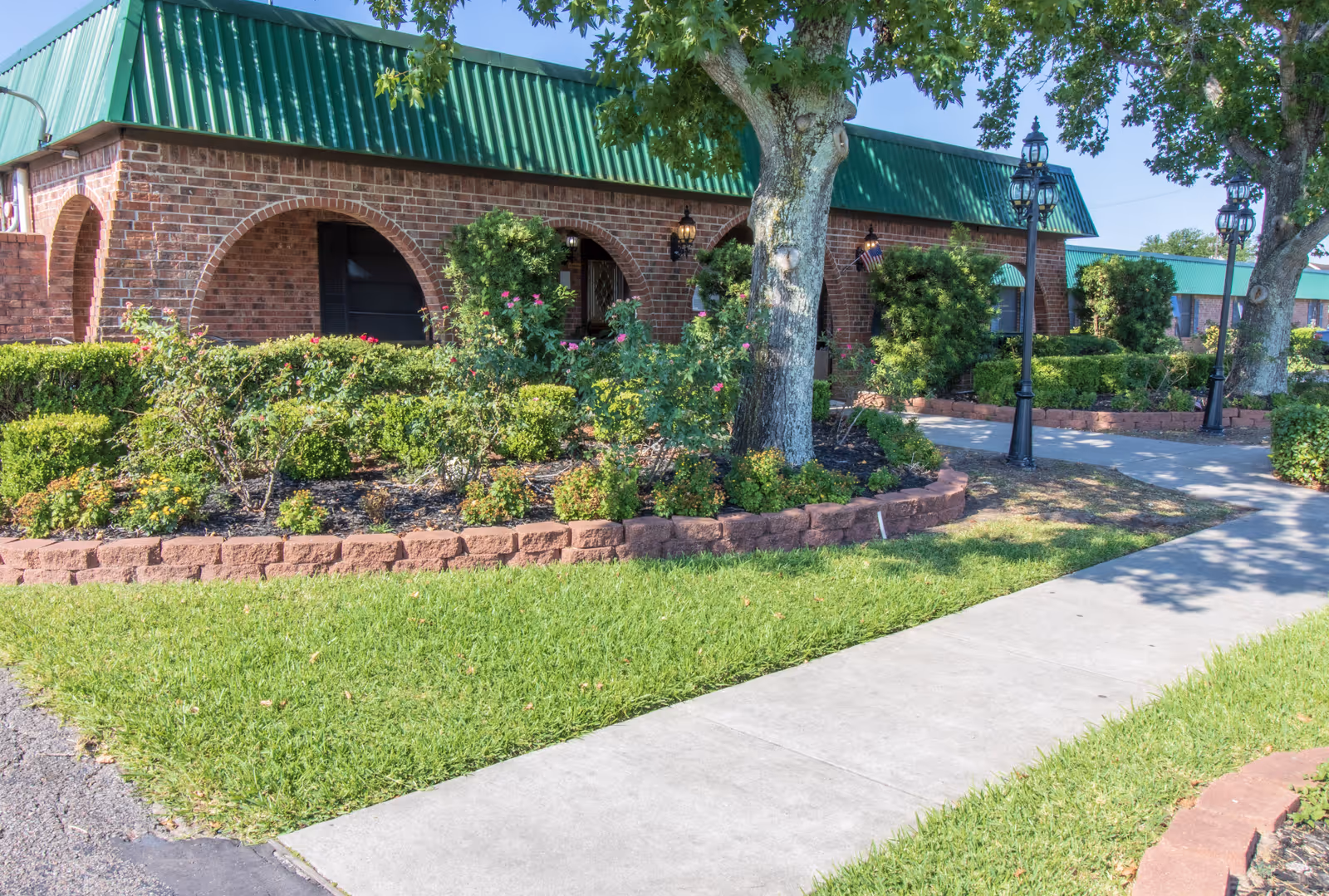 Exterior view of a brick building with a green metal roof, featuring arched doorways and windows. The building is surrounded by well-maintained landscaping including bushes, flowers, and trees. A concrete sidewalk runs alongside the building, and there are black vintage-style street lamps along the path.