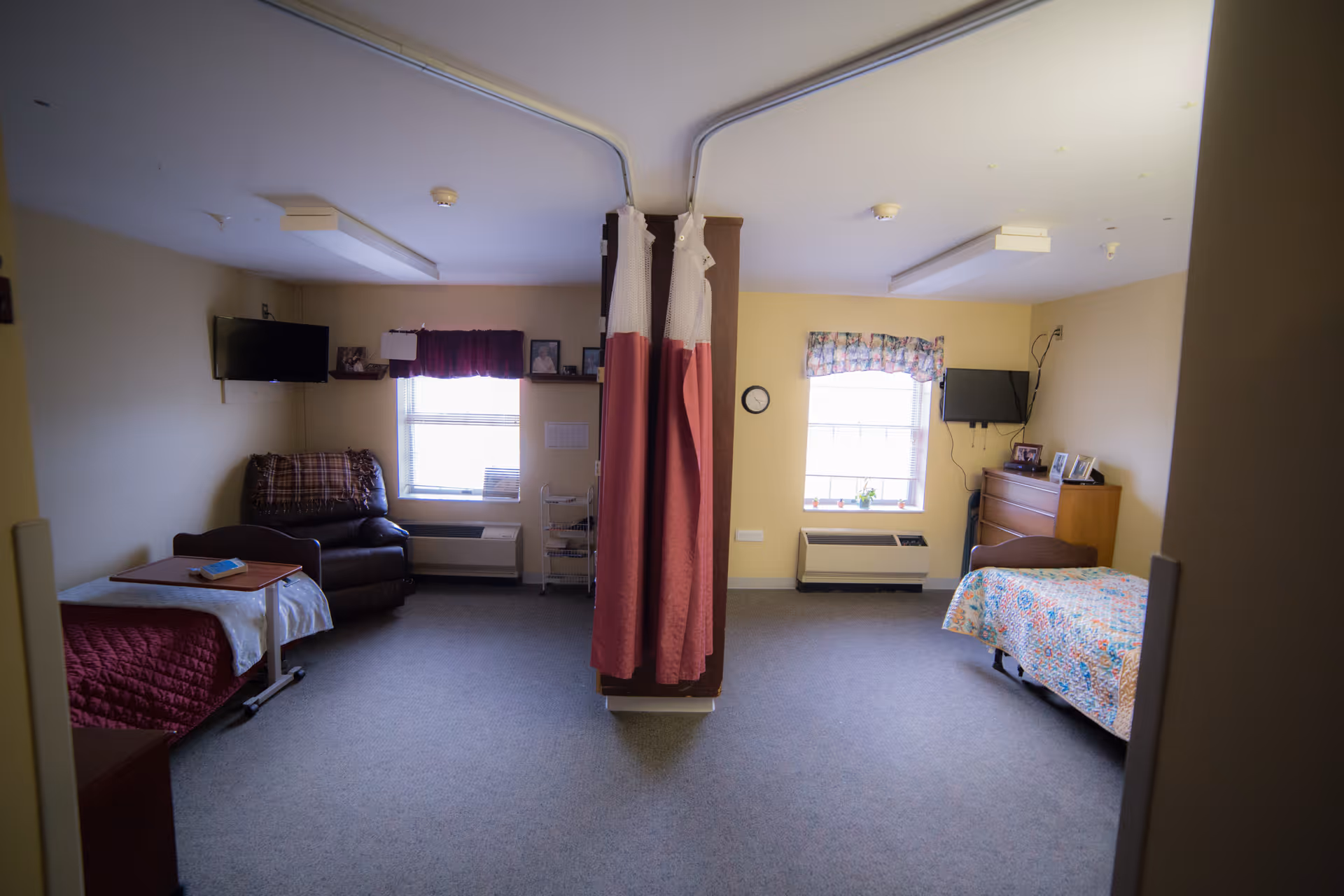 A shared room in a senior living facility with two separate beds divided by a central partition with privacy curtains. Each side has a window with curtains, a wall-mounted TV, and furniture including a chair and a dresser. The room has beige walls and carpeted floor.