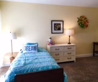 A bedroom with a single bed covered in a blue and white patterned bedspread and a pillow with the word 'DREAM' on it. Next to the bed is a white nightstand with drawers, a lamp, and decorative items. The walls are light-colored with framed artwork and a floral wall decoration. The floor is carpeted.