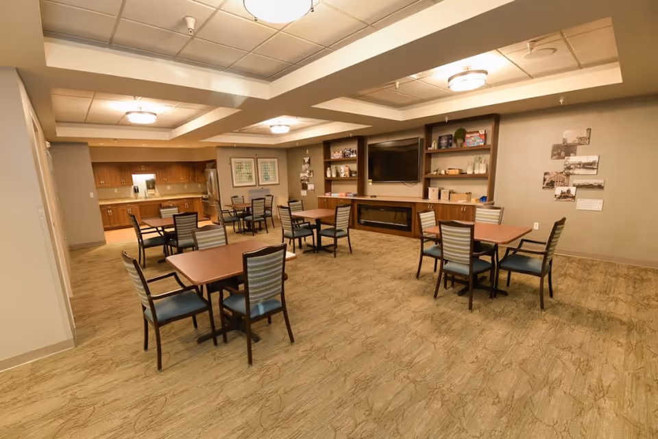 Spacious communal dining and activity room with multiple tables and chairs, a kitchenette and a wall-mounted TV.