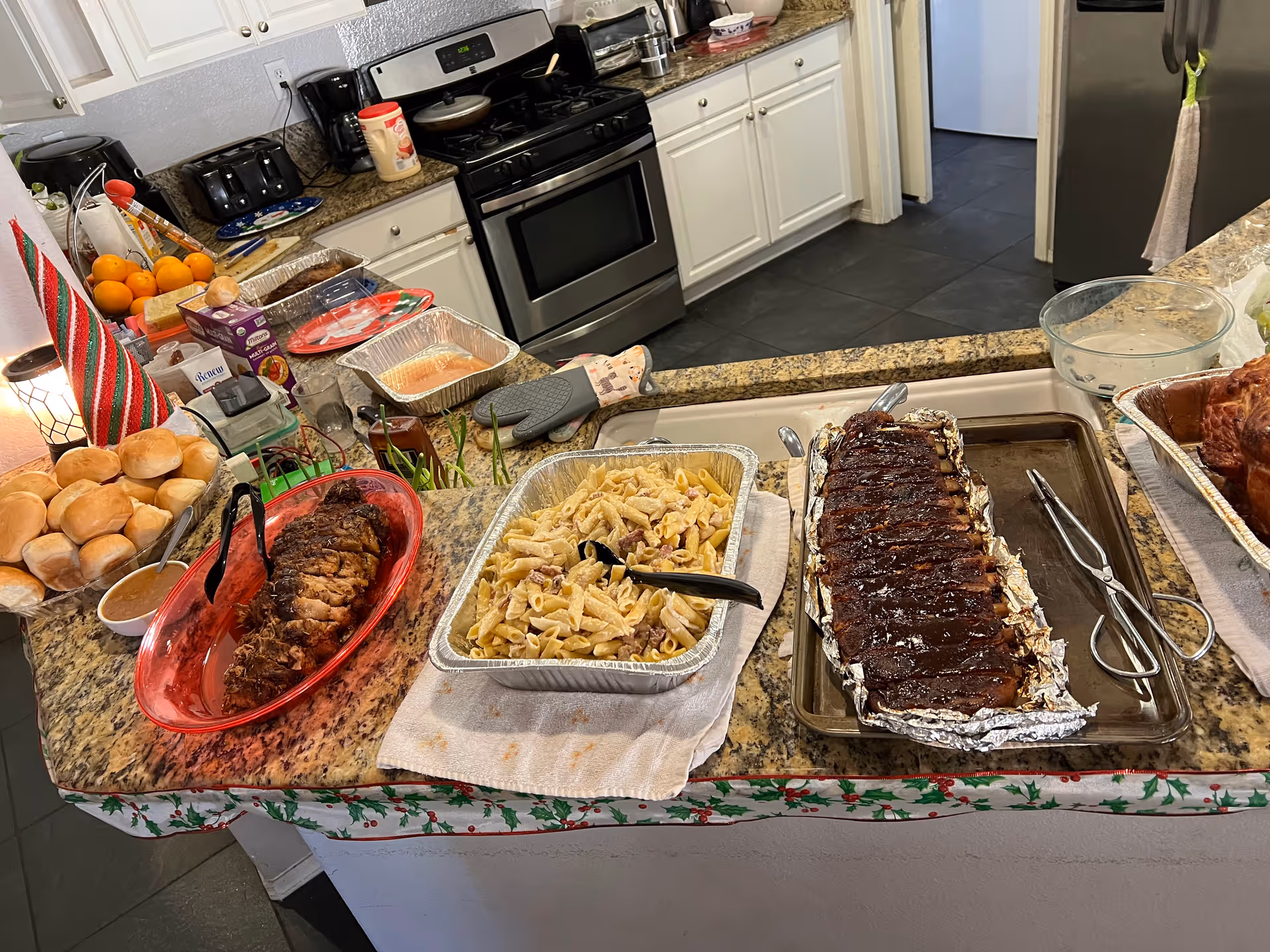 A kitchen counter with a variety of food dishes including a tray of dinner rolls, a plate of sliced roast meat, a tray of pasta salad, and a tray of barbecue ribs. The kitchen has white cabinets, a stainless steel stove, and a refrigerator. Various kitchen items and utensils are visible on the counter and in the background.