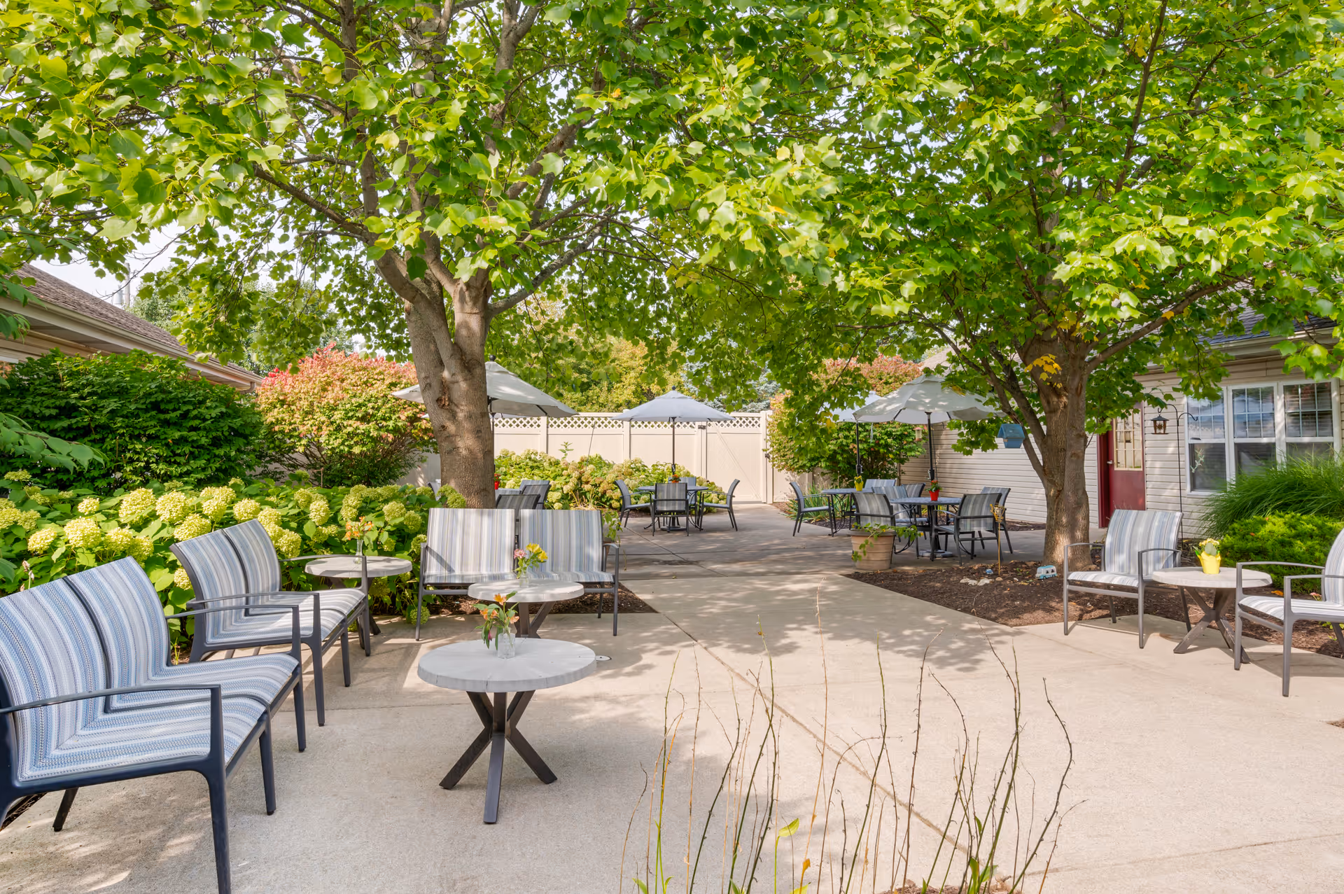 Outdoor patio area with multiple seating arrangements including striped cushioned chairs and small round tables. The space is shaded by large leafy trees and surrounded by green shrubs and flowering plants. Several tables have umbrellas providing additional shade. The area is paved and appears to be part of a senior living facility.