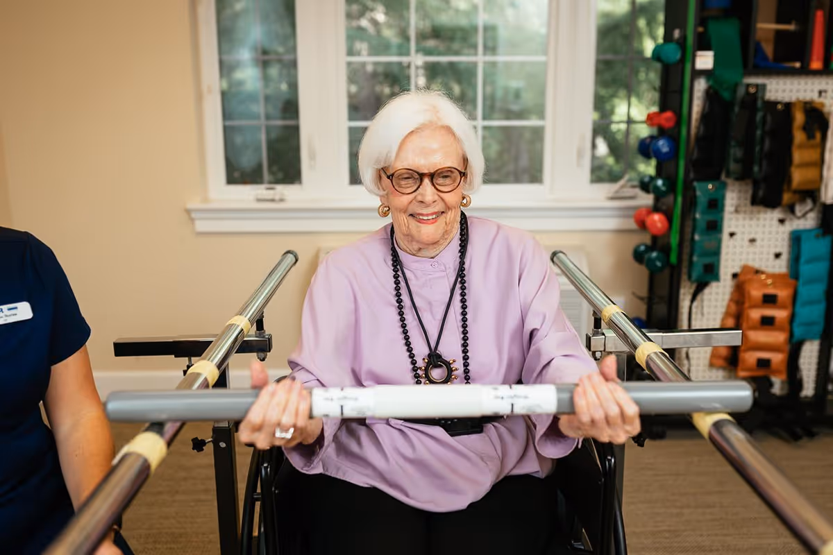 An elderly woman with white hair and glasses is seated in a wheelchair, smiling while holding a gray exercise bar in a rehabilitation or physical therapy room. A person in a navy blue shirt is partially visible beside her. The room has a window with white trim and various exercise equipment, including colorful weights and padded items, on a pegboard wall in the background.