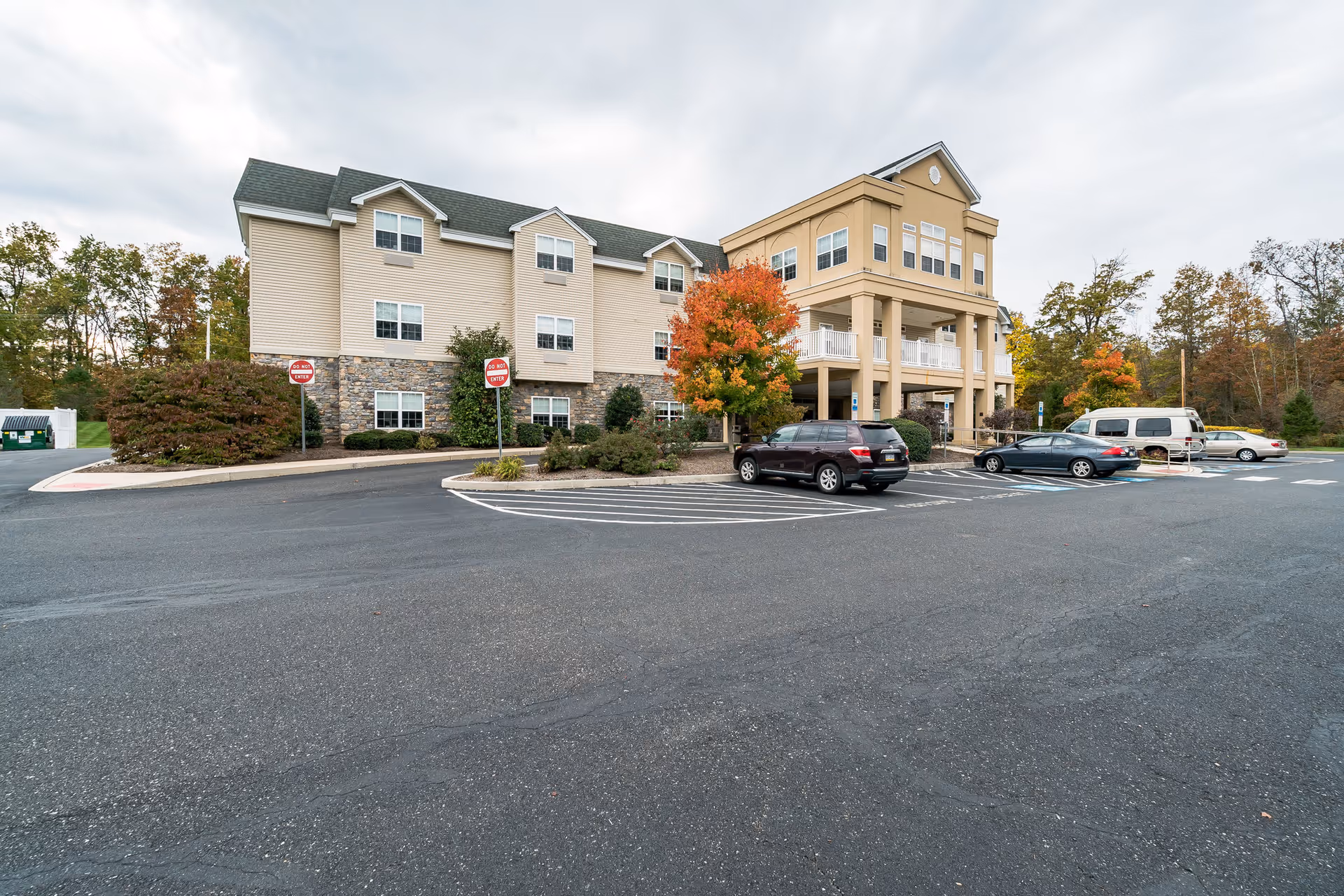 Exterior view of a multi-story senior living facility building with beige siding and stone accents. There are several cars parked in the parking lot in front of the building, and trees with autumn foliage surround the area under a cloudy sky.