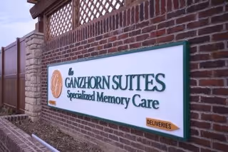 A large outdoor sign mounted on a brick wall with lattice fencing above it. The sign reads 'The Ganzhorn Suites Specialized Memory Care' with an orange circle logo on the left and a small orange arrow labeled 'Deliveries' on the bottom right.