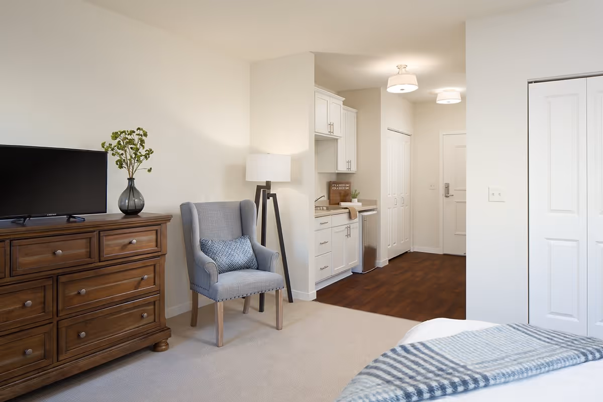 A cozy interior space featuring a wooden dresser with a flat-screen TV and a vase with greenery on top, a gray upholstered armchair with a blue patterned pillow, a floor lamp, and a small kitchenette area with white cabinets, a sink, and a mini refrigerator. The room has light-colored walls and carpeted flooring transitioning to wood flooring near the kitchenette and entrance door.
