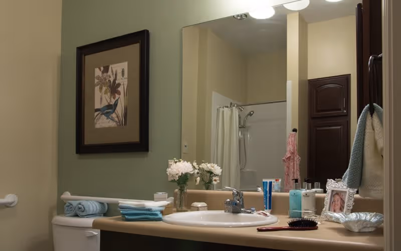 Bathroom sink area with a large mirror above it. On the countertop are a vase with white flowers, folded towels, toiletries, a hairbrush, and a framed photo. A shower with a white curtain and a dark wooden cabinet are visible in the background. A framed picture of a bird hangs on the wall above the toilet.