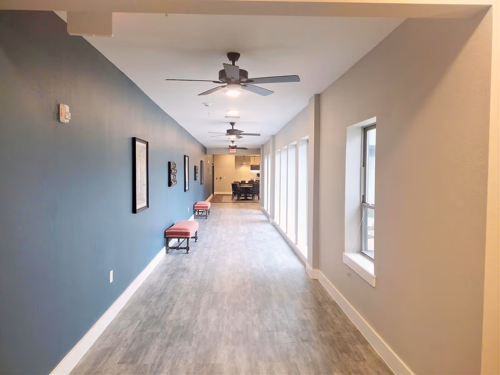 A long hallway with a blue accent wall on the left and large windows on the right. The hallway has ceiling fans with lights, framed artwork on the blue wall, and two red cushioned benches. At the end of the hallway, there is a dining area with a table and chairs.