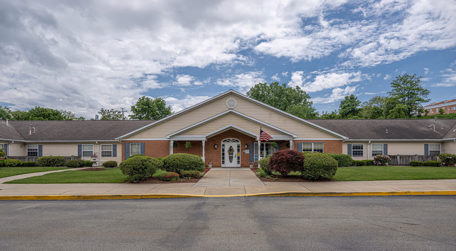 Front exterior view of Arden Courts - ProMedica Memory Care Community in Monroeville, showing a single-story building with a peaked roof, beige siding, brick accents, an American flag near the entrance, and well-maintained landscaping with bushes and a sidewalk leading to the front door under a partly cloudy sky.