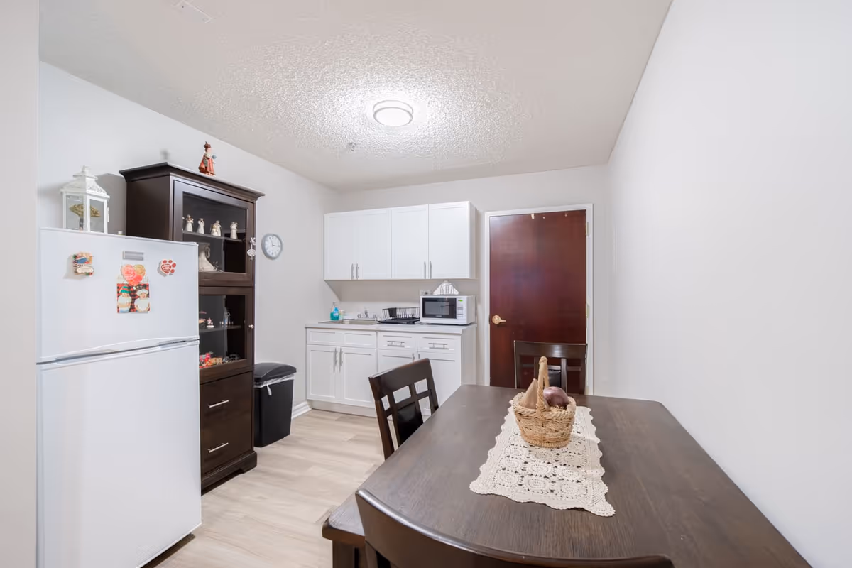 A small kitchen and dining area with a dark wooden table and chairs. The table has a lace runner and a basket with fruit. The kitchen area has white cabinets, a microwave, a dish rack, and a white refrigerator with magnets. There is a dark wooden cabinet next to the refrigerator and a wall clock above a trash bin. The walls are white and the floor is light wood.