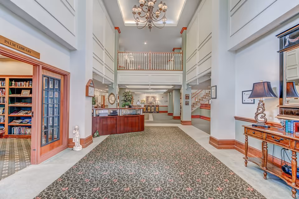 Spacious and well-lit interior lobby area of a senior living facility with a patterned carpet, chandelier, wooden reception desk, and a library room labeled 'Palm Library' with bookshelves visible through glass doors. There is a wooden table with a lamp and mirror on the right side, and a staircase leading to an upper level in the background.