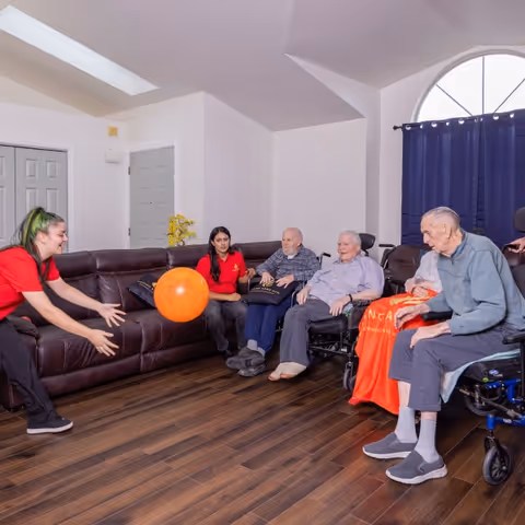 A group of elderly people and two caregivers in a living room. The caregivers, wearing red shirts, are playing with an orange ball with the elderly individuals, some of whom are seated in wheelchairs. The room has wooden flooring, a large brown leather sectional sofa, and a window with a dark curtain.
