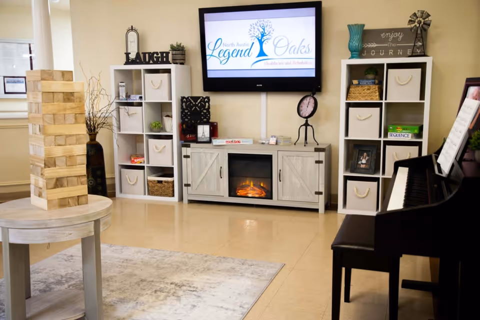 A cozy common area with a large wooden Jenga game on a round table, two white shelving units with decorative items and storage baskets, a wall-mounted TV displaying the Legend Oaks Healthcare and Rehabilitation - North Austin logo, an electric fireplace below the TV, and a black piano with sheet music on the right side.