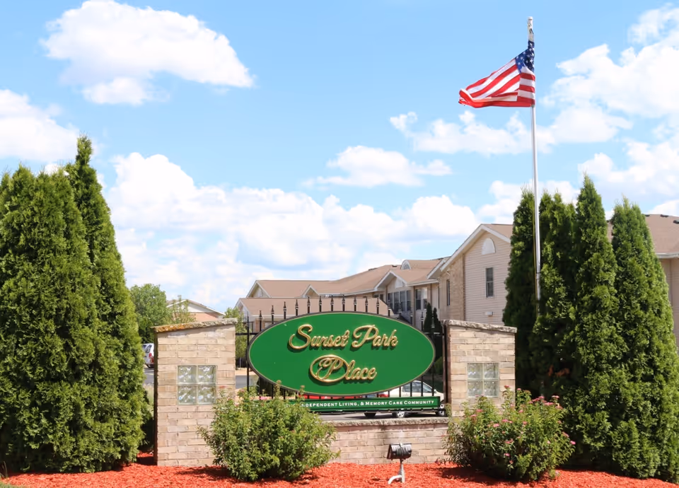 Entrance sign for Sunset Park Place, a senior living community, displayed on a green oval board mounted on a brick structure with bushes and tall evergreen trees on either side. An American flag is flying on a flagpole to the right, with residential buildings visible in the background under a partly cloudy blue sky.