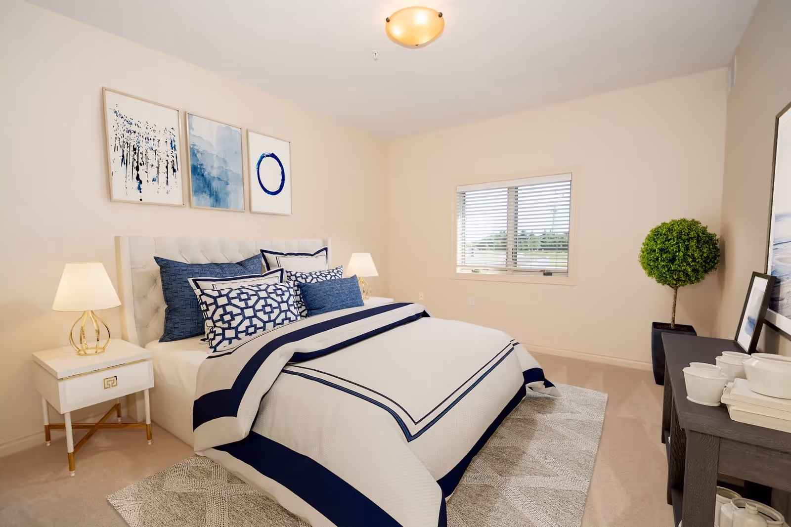 A neatly made bed with white and navy blue bedding in a bedroom. The bed has multiple decorative pillows and a tufted headboard. On the left side, there is a white nightstand with a lamp. Above the bed, three abstract blue and white paintings are hung on the beige wall. A window with blinds is on the right wall, and a potted green plant stands next to a dark wooden console table with white ceramic dishes and books on it. The room has beige carpet and a ceiling light fixture.