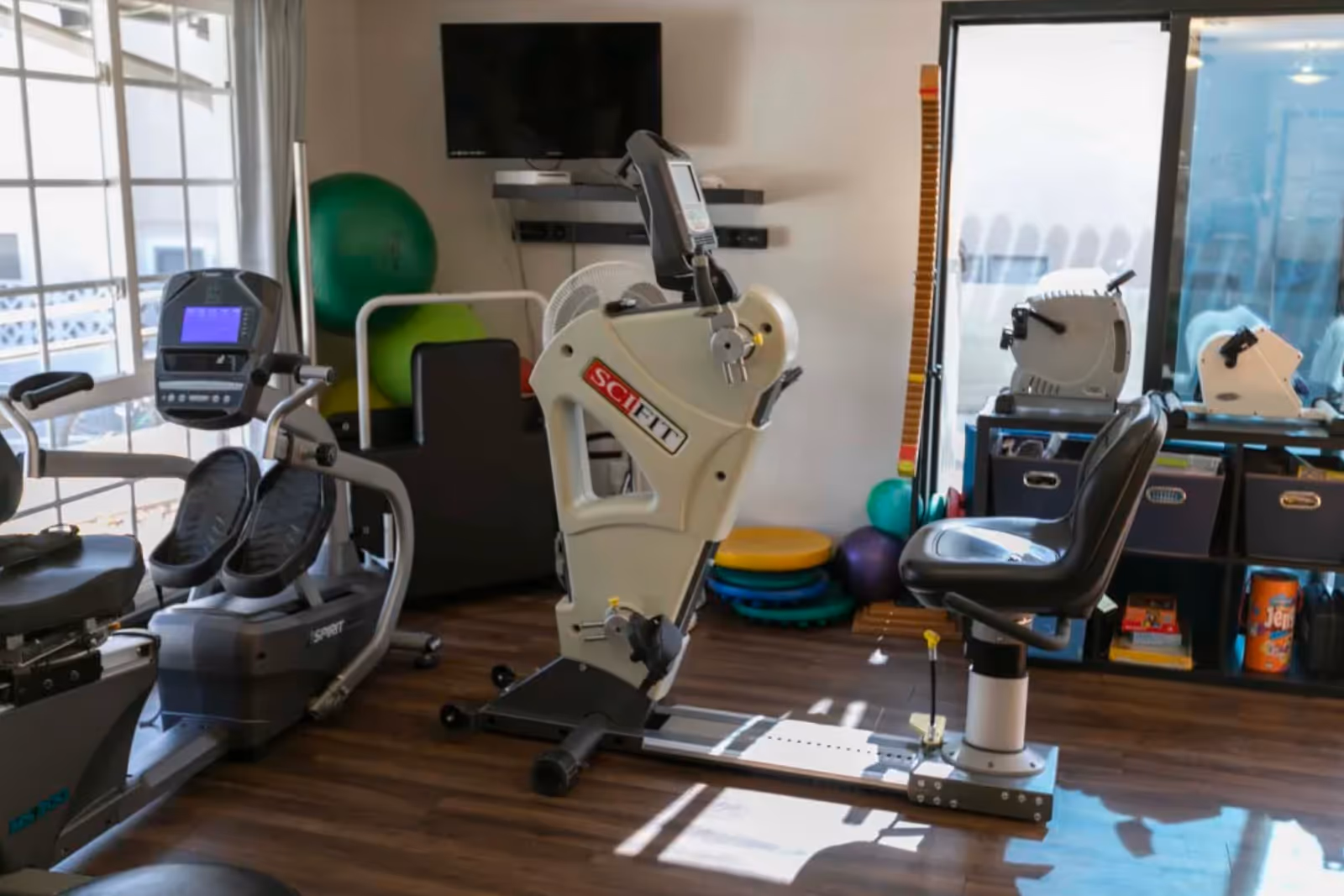 A well-lit exercise room with various fitness equipment including a recumbent bike, a SciFit exercise machine, and other workout devices. There are large windows letting in natural light, a TV mounted on the wall, and shelves with storage bins and exercise balls in the background.