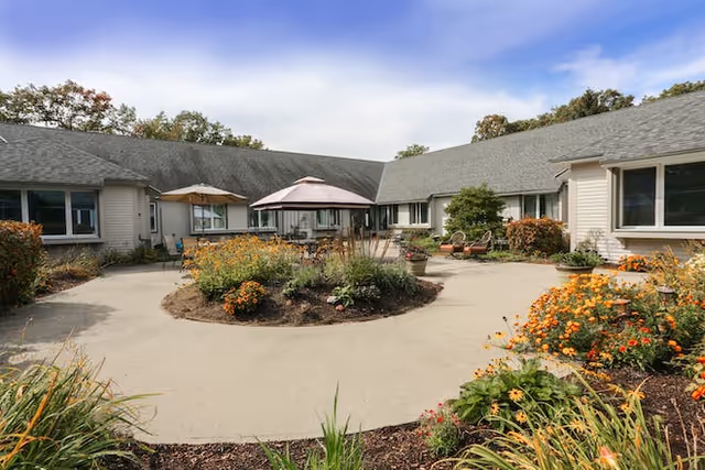 A landscaped circular courtyard with patio tables, umbrellas, and flowering beds surrounded by single-story senior living buildings.