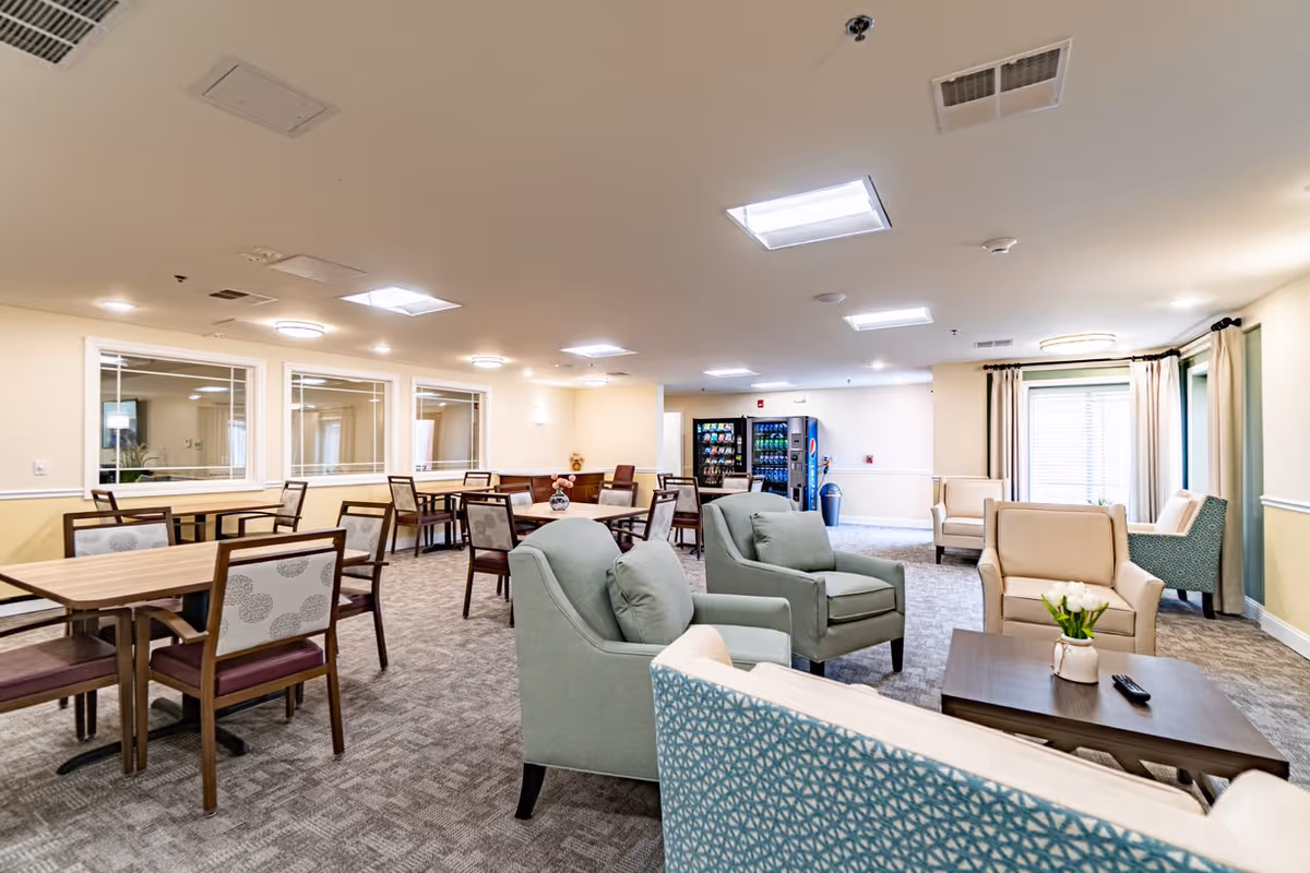 A spacious and well-lit common area in a senior living facility featuring multiple seating arrangements including armchairs and tables with chairs. The room has carpeted floors, large windows with curtains, and vending machines in the background. The walls are painted in light colors, and the ceiling has several recessed lights.