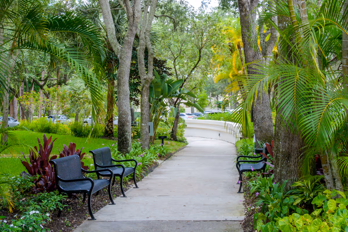 A paved walkway lined with black metal benches on both sides, surrounded by lush green trees, plants, and shrubs in a garden setting.