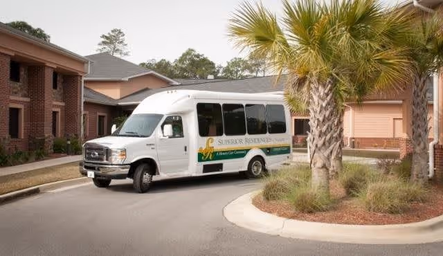 A white shuttle bus with the logo and text 'Superior Residences' parked on a curved driveway in front of a brick building with palm trees and landscaping.
