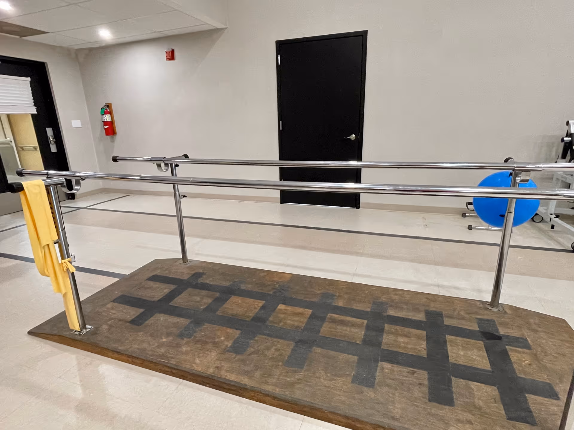 Indoor physical therapy area with parallel metal handrails mounted on a wooden platform with black tape markings. A yellow towel hangs on the left handrail. In the background, there is a black door, a fire extinguisher mounted on the wall, and a blue exercise ball on the right side.