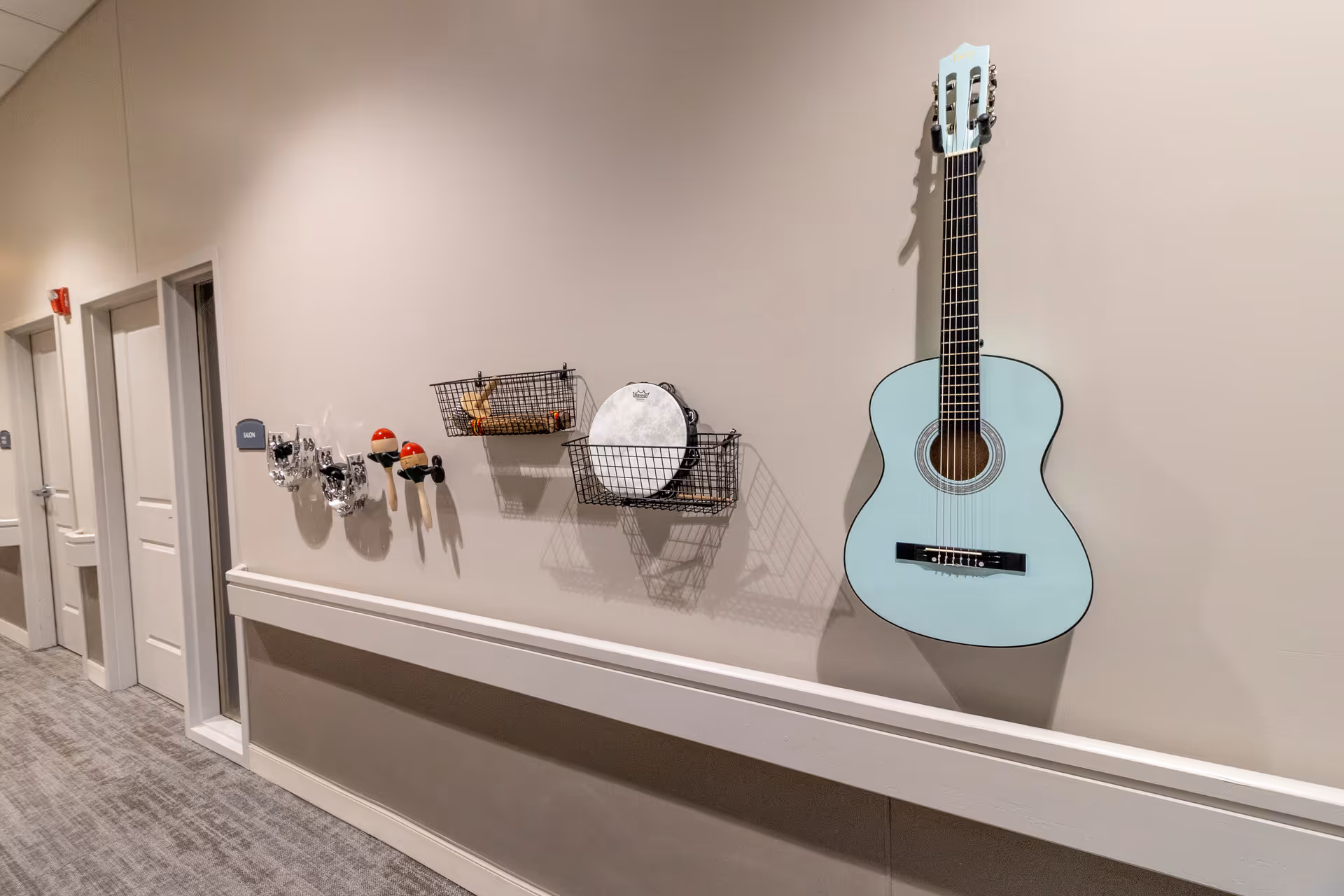 A hallway with light-colored walls and carpeted floor featuring a mounted light blue acoustic guitar, two wire baskets holding percussion instruments including a tambourine and maracas, and several other percussion instruments mounted on the wall. There are three closed doors along the hallway, one labeled 'Salon'.