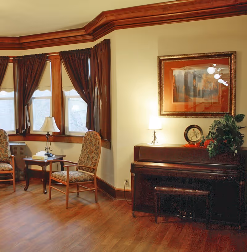 Cozy seating area with two upholstered chairs by bay windows, a side table with a lamp, and a piano topped with a lamp and framed artwork.