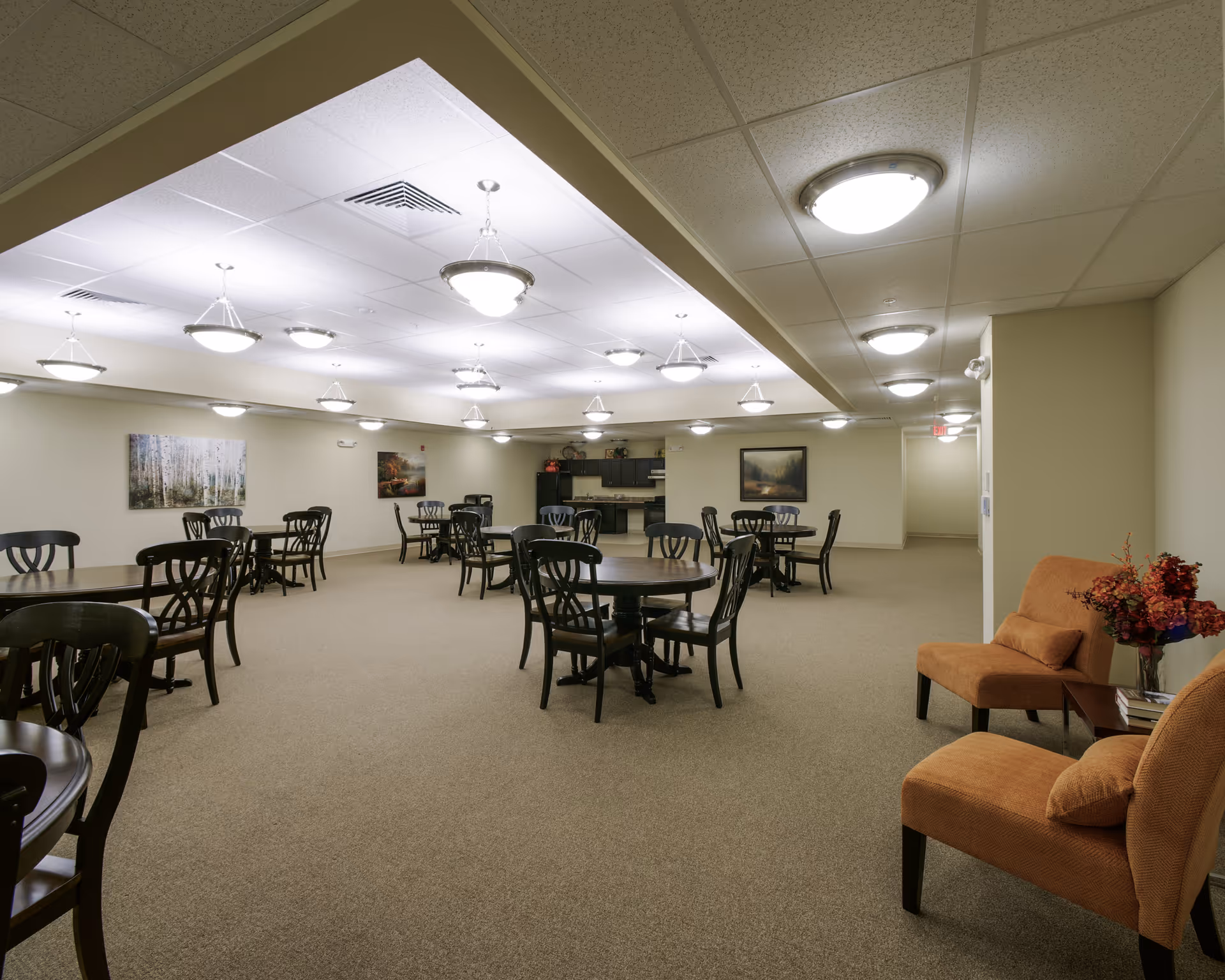 A spacious dining room with multiple round wooden tables and black chairs arranged neatly. The room has beige walls and carpet, with several ceiling lights providing bright illumination. There are two orange upholstered chairs with cushions and a small table with a flower arrangement on the right side. Paintings hang on the walls, and a kitchenette area is visible in the background.