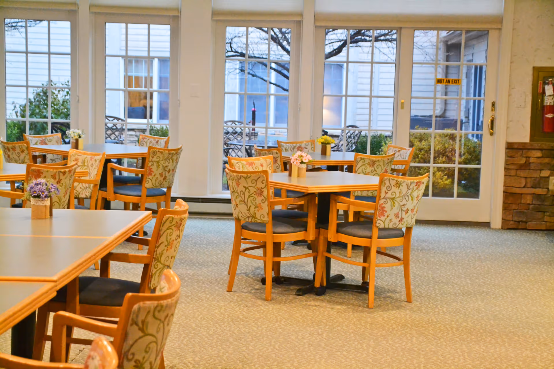 A bright dining room with several wooden tables and chairs featuring floral patterned upholstery. Small flower arrangements are placed on each table. Large windows and glass doors provide a view of an outdoor patio area with additional seating and greenery.