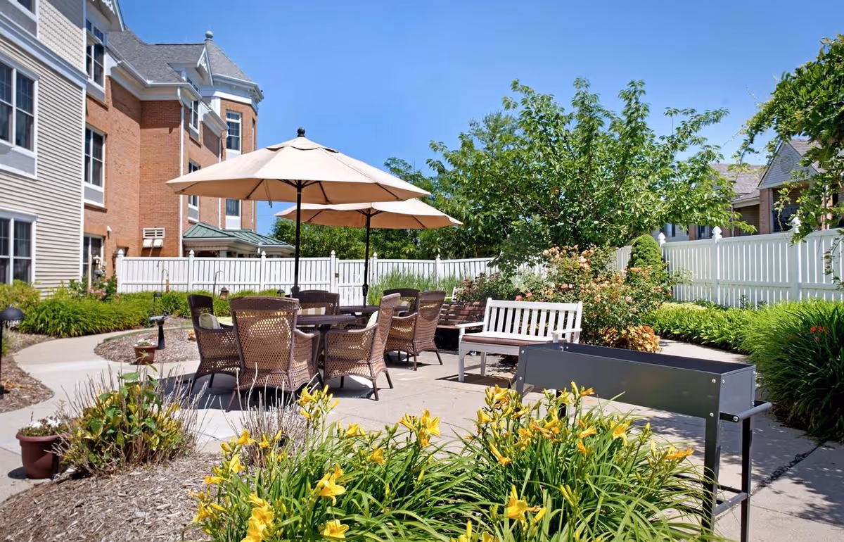 Outdoor patio area at a senior living facility with a round table surrounded by wicker chairs under two large beige umbrellas. There is a white wooden bench, a black metal planter, and various green plants and yellow flowers around the paved patio. The building and a white fence are visible in the background under a clear blue sky.
