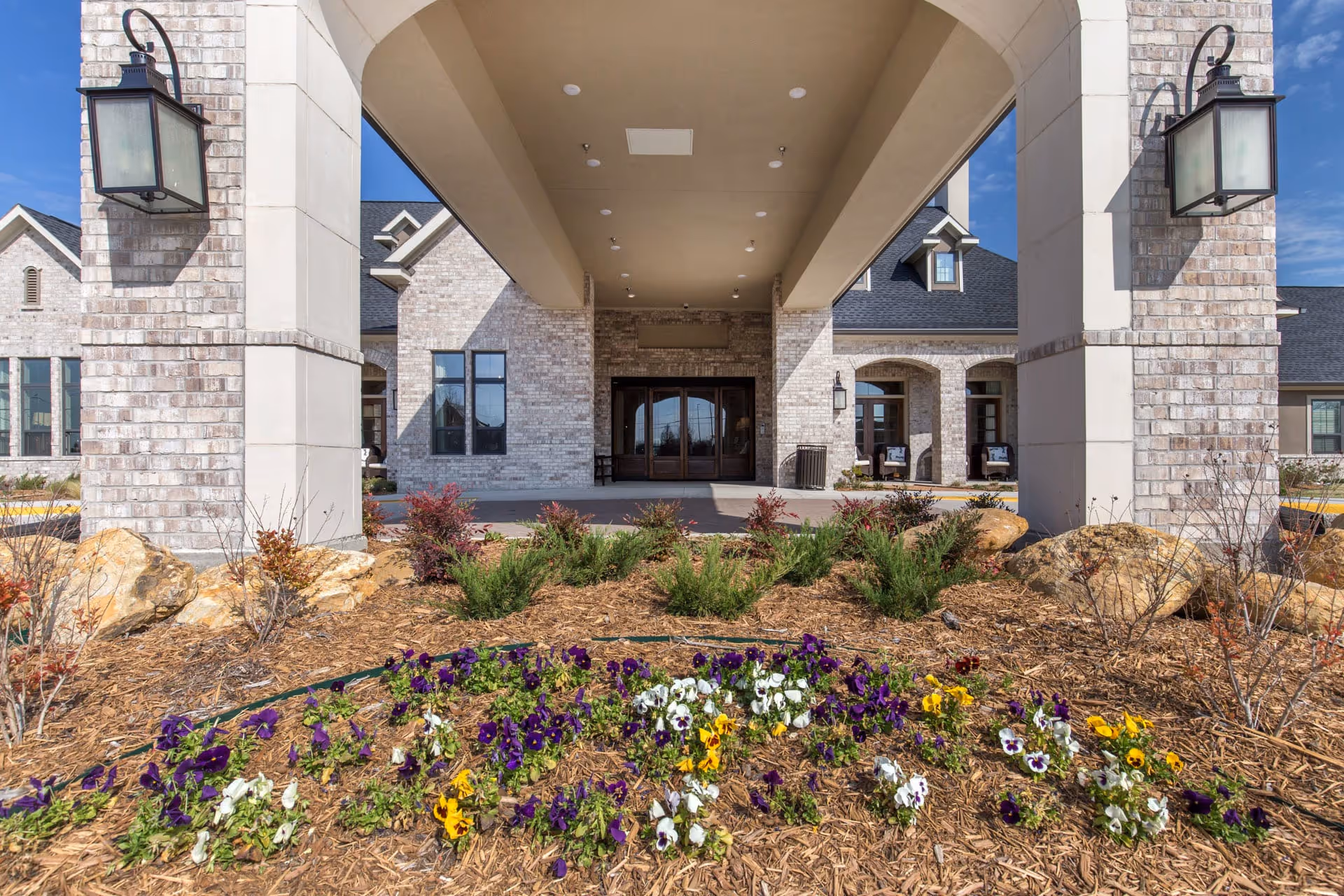 Front entrance of a brick senior living building with a covered porte-cochere, flowerbeds, and large lanterns.