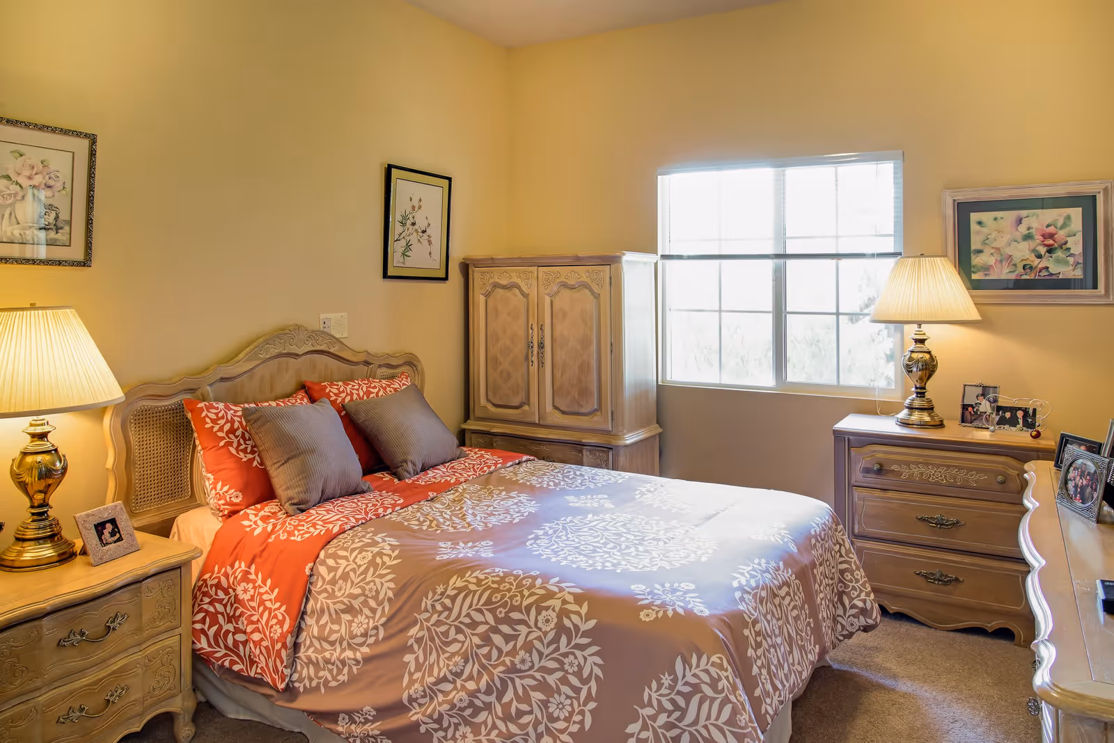 A cozy bedroom with a bed featuring a red and beige floral patterned comforter and pillows. The room has two wooden nightstands with lamps, a wooden dresser, and a cabinet. There are framed floral artworks on the walls and a window letting in natural light.