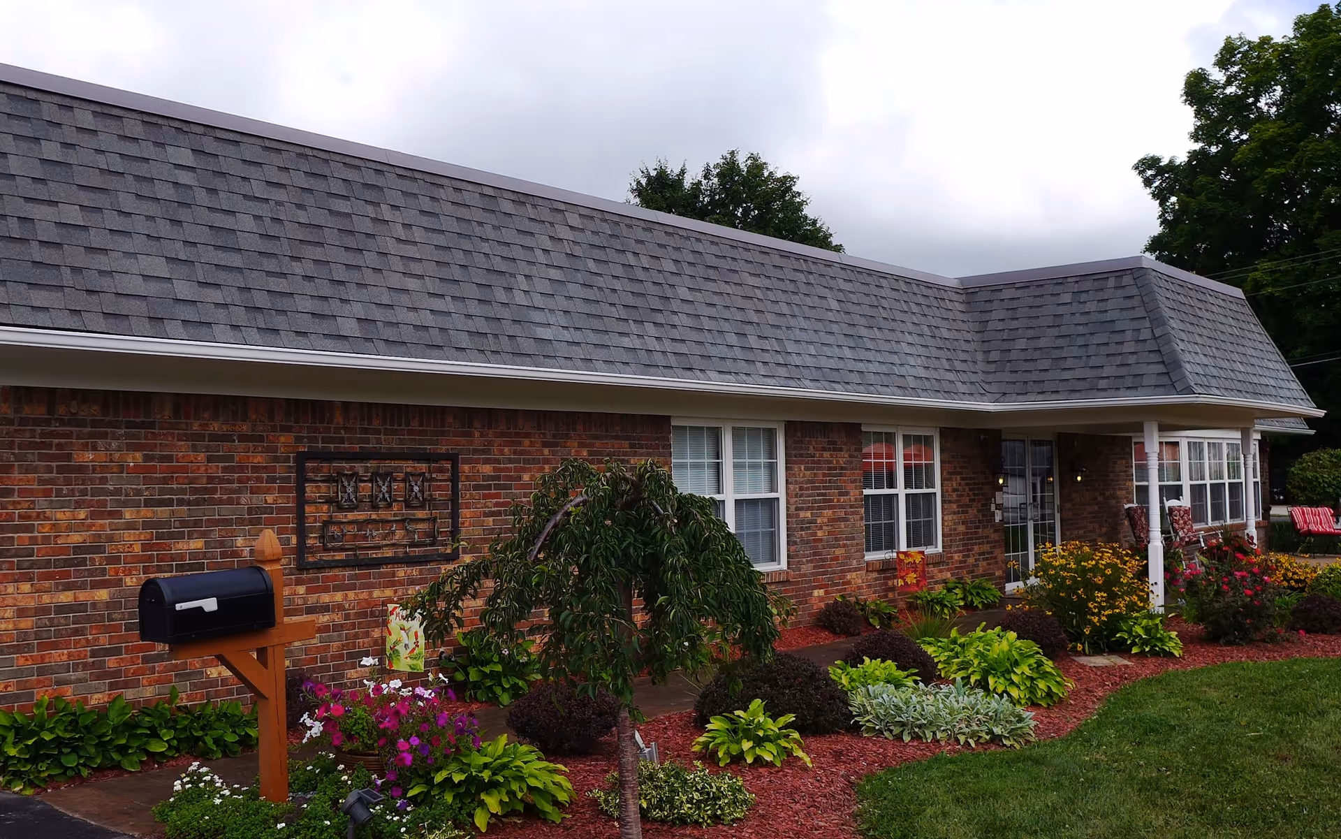 Exterior view of a single-story brick building with a gray shingled roof, surrounded by a well-maintained garden with various green plants and colorful flowers. There is a black mailbox on a wooden post in the foreground and a covered entrance with glass doors and windows.