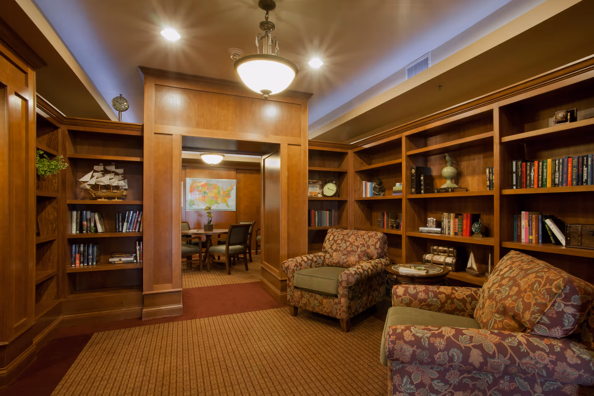 A cozy library or reading room with wooden bookshelves filled with books and decorative items. Two upholstered armchairs with floral patterns are positioned near a small round table. The room has warm lighting from a ceiling fixture and recessed lights. Through a wooden doorway, a conference or meeting room with a round table and chairs is visible, along with a colorful map on the wall.