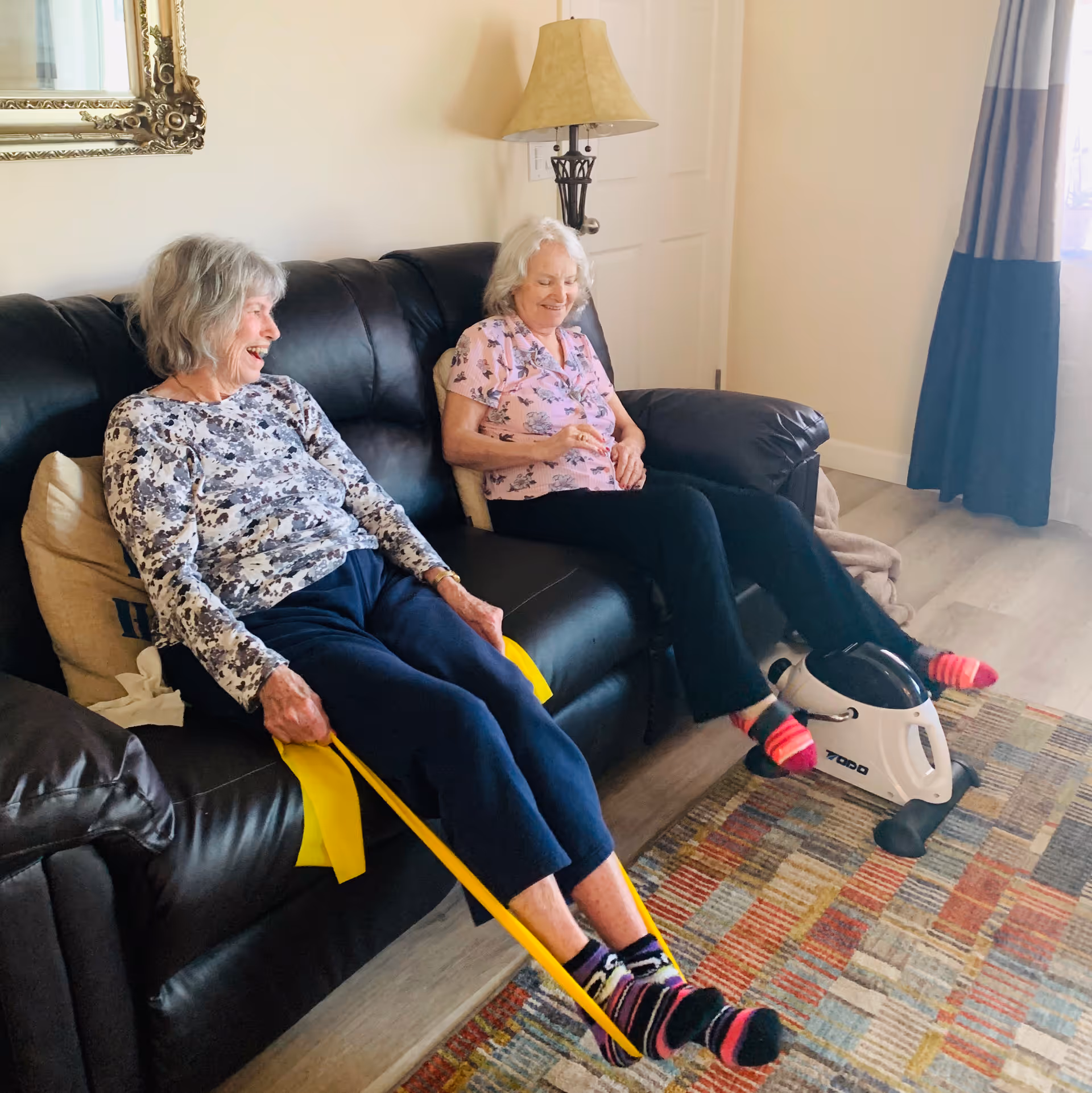 Two elderly women sitting on a black leather couch in a living room. One woman is using a yellow resistance band for leg exercises, while the other is using a small pedal exerciser. They both appear happy and engaged in light exercise. The room has a patterned rug, a lamp, a large mirror on the wall, and a window with curtains.
