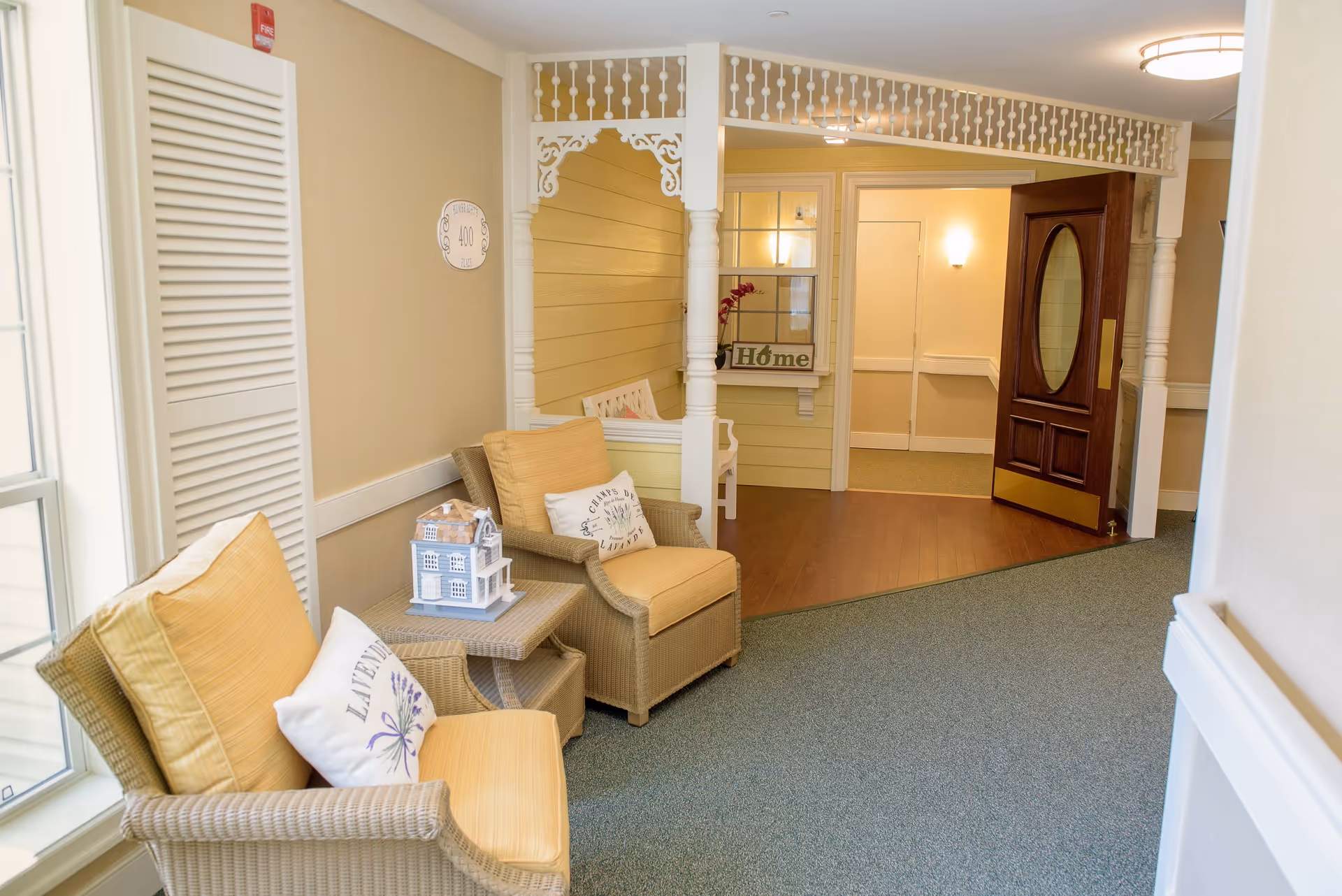 A cozy seating area in a senior living facility hallway with two wicker armchairs featuring yellow cushions and decorative pillows. A small table between the chairs holds a miniature house decoration. The hallway has a carpeted floor, a wooden door with an oval glass window, and a decorative white railing above a yellow paneled wall with a window and a 'Home' sign.