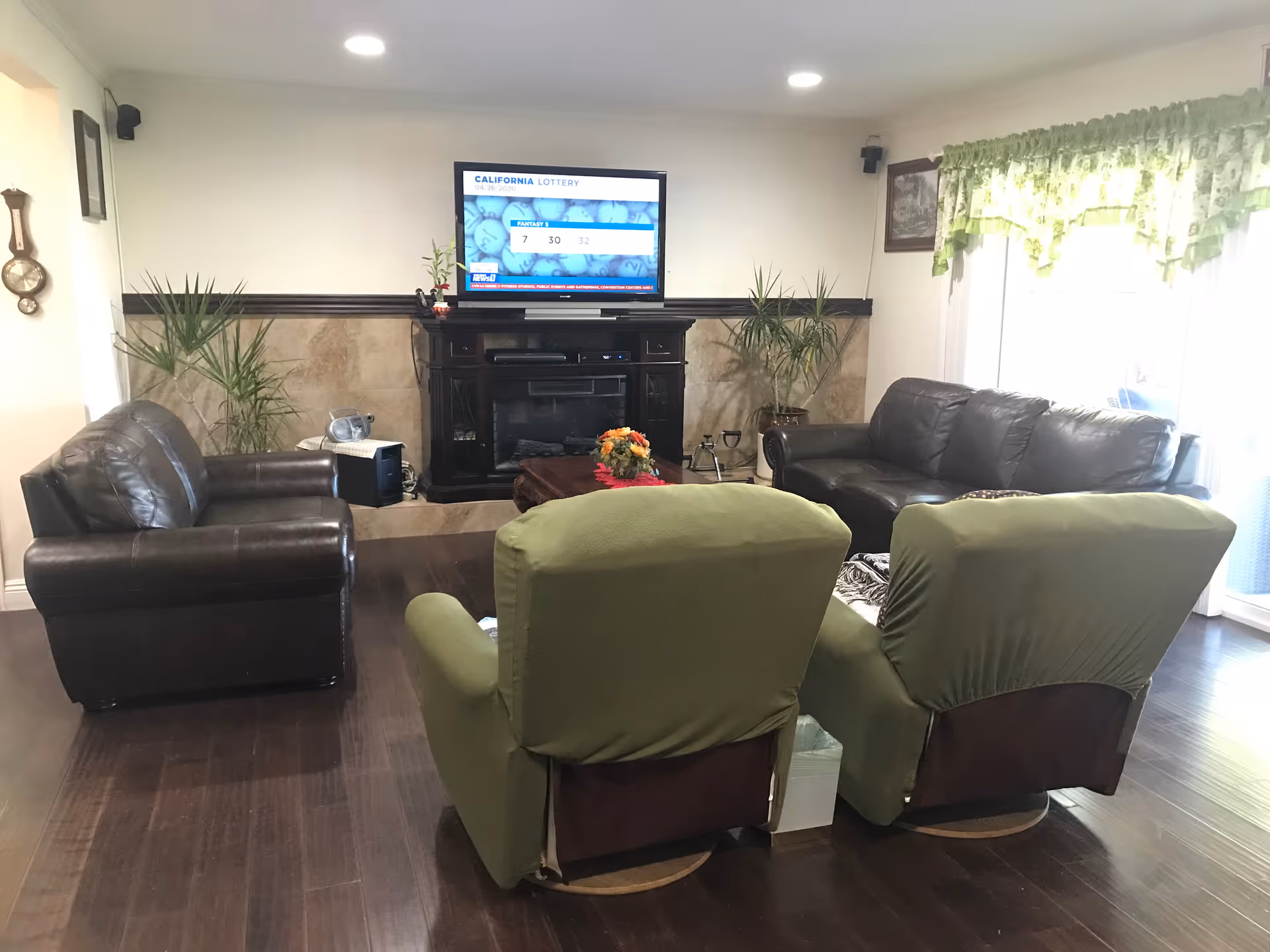 A cozy living room with two green armchairs in the foreground, a dark brown leather sofa and loveseat, a wooden coffee table with a flower arrangement, two potted plants flanking a fireplace with a mounted flat-screen TV displaying a California Lottery screen, and a window with green floral curtains on the right.