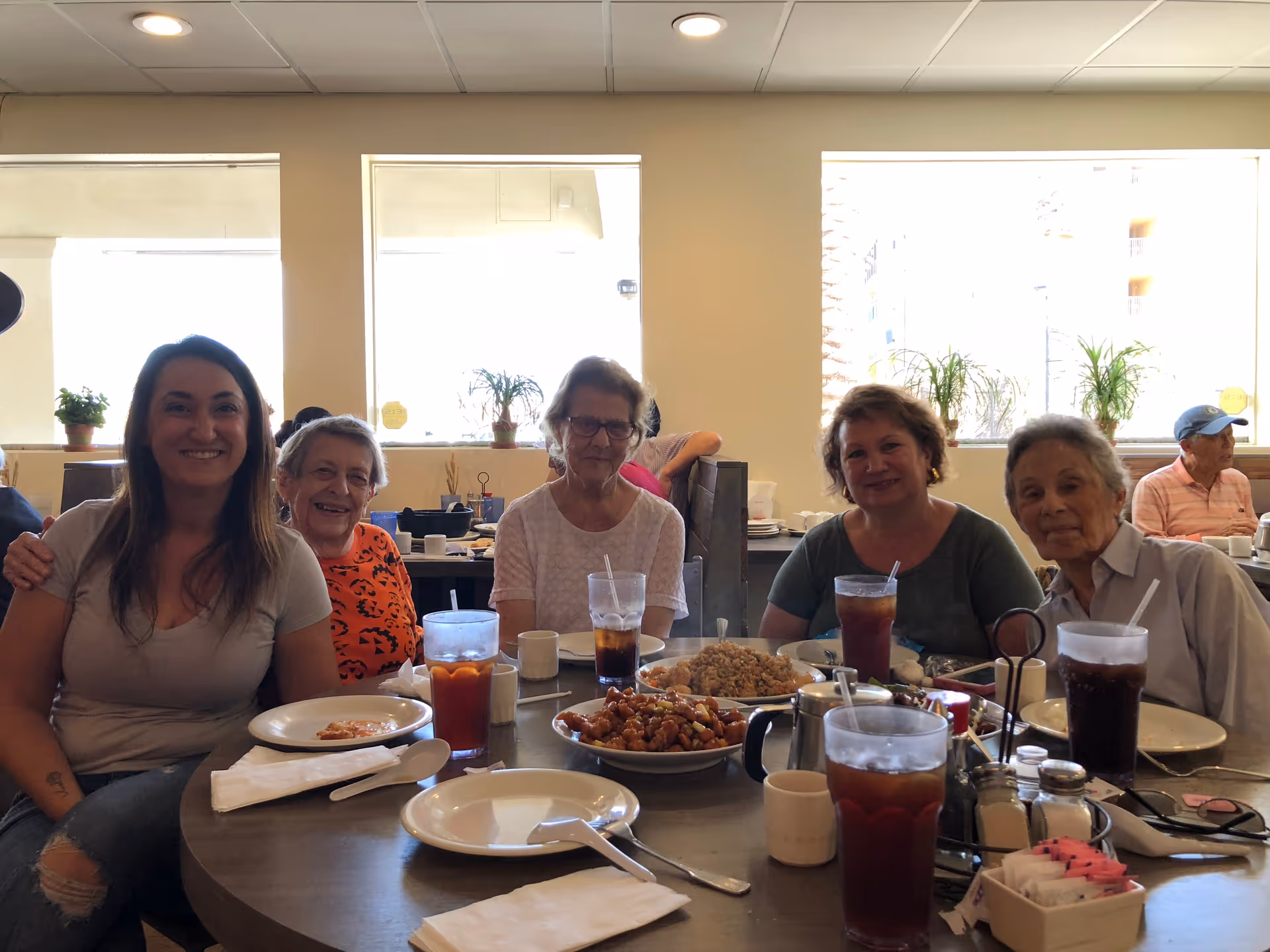 Five women seated around a table in a bright dining room with plates, bowls of food, and glasses of iced tea.