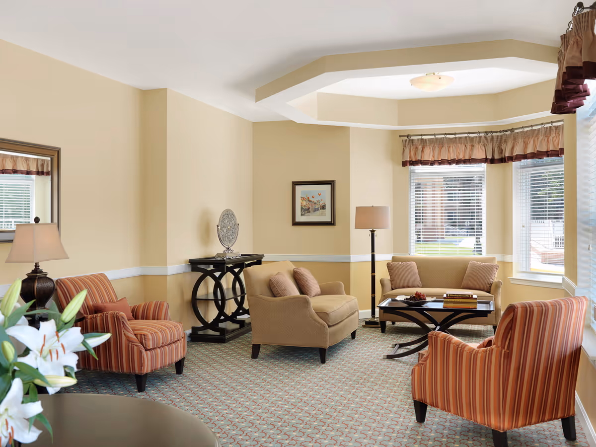 A bright, carpeted living room with striped armchairs, a loveseat, coffee table, and large windows.